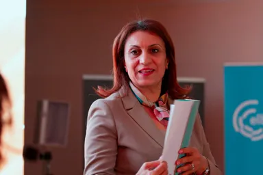 A woman in a beige blazer with a patterned scarf smiles while holding white and green folders, standing in front of a blurred blue banner with a partly visible logo.
