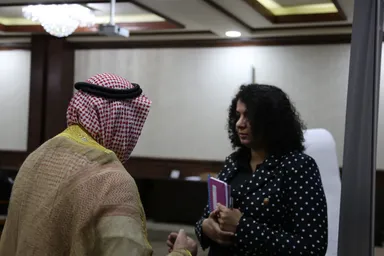 A man wearing a checkered keffiyeh converses with a woman holding a folder in a meeting hall with a chandelier.