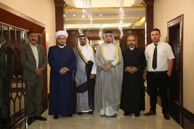 A group of men wearing a mix of suits, turbans and traditional robes pose together in a decorated hallway with a glass door and hanging lights.