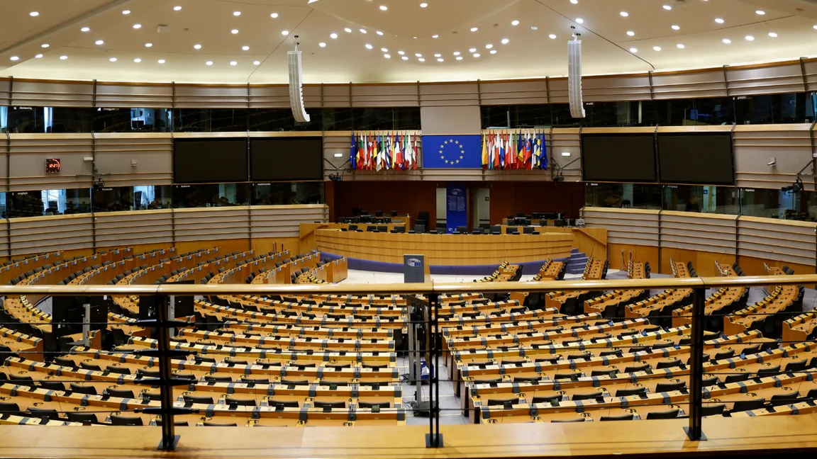 An empty parliamentary chamber with curved rows of desks and monitors, flags including the EU flag at the front beneath a paneled ceiling.