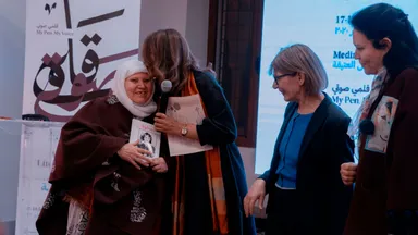 A woman wearing a white headscarf embraces another woman on stage while two other women look on, near a banner reading "My Pen. My Voice.".