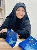 A woman in a headscarf sits at a table holding a blue book, with more copies of the same book beside her.