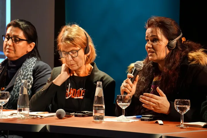 Three women sit at a panel table, one speaking into a microphone while wearing headphones, with water glasses in front of them.