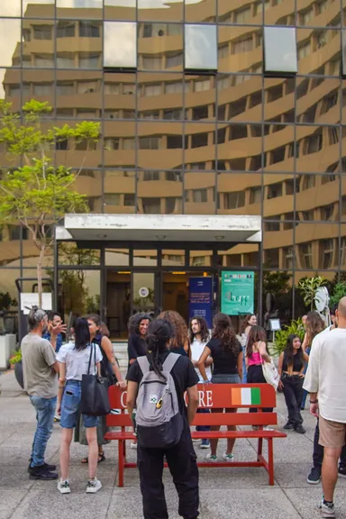 A group of people standing outside a glass-fronted building, near a red bench painted with a flag design and partly legible lettering.
