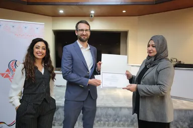 A man in a suit hands a certificate to a woman in a headscarf while another woman stands smiling beside them, in front of a banner with Arabic text.
