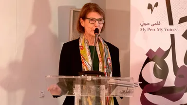 A woman speaking into a microphone at a lectern, beside a banner reading "My Pen. My Voice." with Arabic text and calligraphy.