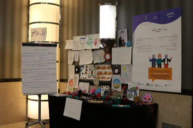 A display of children's drawings, handwritten notes and craft items stands beside a poster reading "You Can" and "بتقدري", showing an illustration of three women.