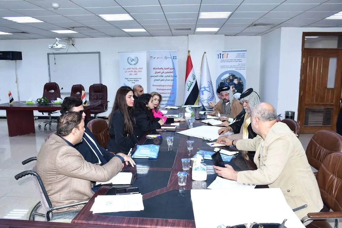 People sit around a conference table in a meeting room with banners reading "Promoting Inclusive Dialogue and Participation in Iraq", flags, and a man in a wheelchair in the foreground.