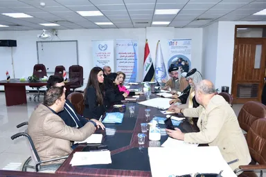 People sit around a conference table in a meeting room with banners reading "Promoting Inclusive Dialogue and Participation in Iraq", flags, and a man in a wheelchair in the foreground.