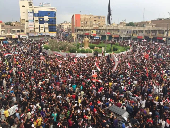 Aerial view of a densely packed crowd holding flags in a city square lined with buildings and shops, with two statues on a central traffic island.