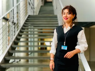 A woman stands on a staircase landing wearing a black dress with a leopard-print collar and a visitor badge on a lanyard.