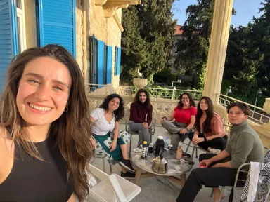 A woman takes a selfie with five people seated around a table on a balcony surrounded by trees.