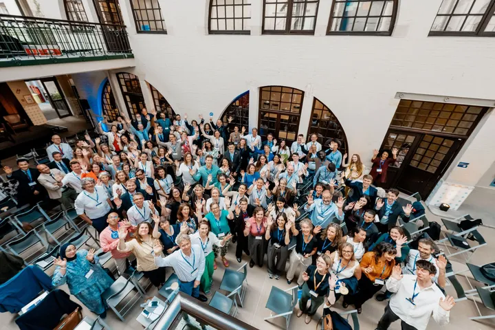 An aerial view of a large group of people in a courtyard waving up at the camera, wearing lanyards.