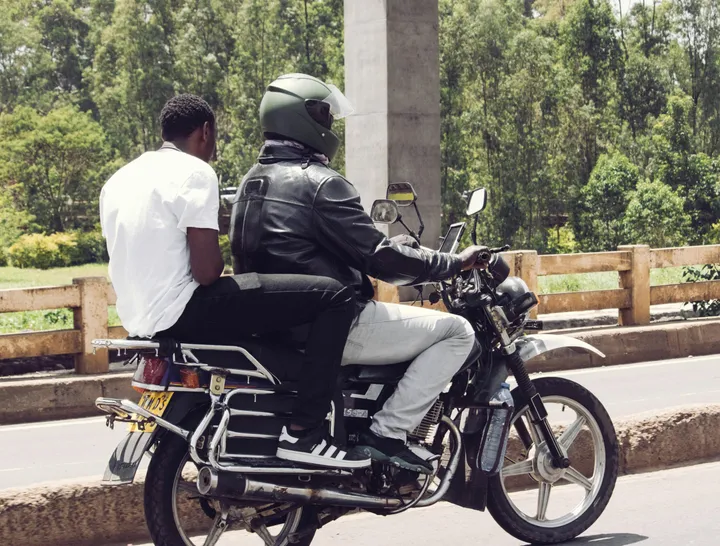 Two men ride a motorcycle along a road lined with trees and a wooden fence.
