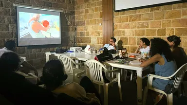 A group of people sit at tables watching an animated video projected on a screen showing a cartoon boxer.