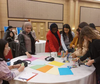 A group of women gather around a round table covered with colored paper sheets, markers and craft supplies in a conference hall.
