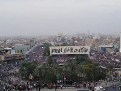 Aerial view of a large crowd filling a plaza with palm trees, overlooked by a banner showing silhouetted figures, against a hazy city skyline.