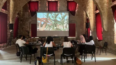 People seated at tables in a vaulted stone hall with red curtains, watching a projected image of plants on a screen.