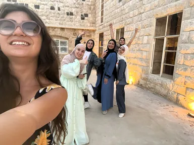 Selfie showing a woman in sunglasses in the foreground, with a group of women posing and jumping behind her outside a stone building.