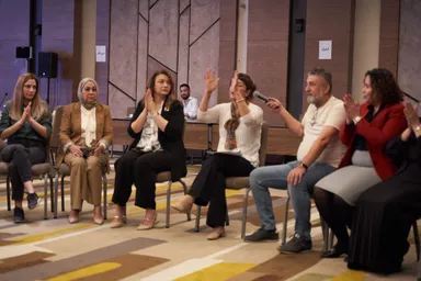 A group of people sits on chairs, several applauding or raising a hand, one holding a microphone, with signs reading "ذي قار" and "أربيل" on the wall behind them.