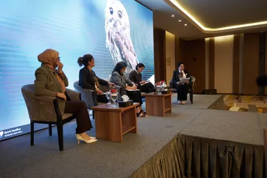 Five people sit in armchairs on a stage in front of a large screen showing an owl image and a news ticker, with water glasses on small tables between them.