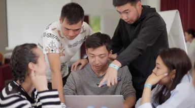 A group of five people gathered around a laptop, with one man pointing at the screen.