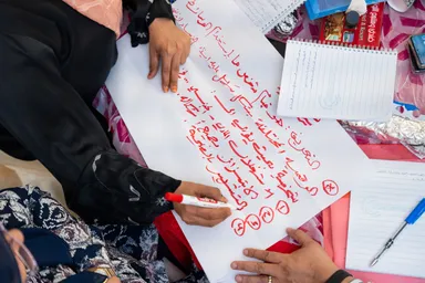 Hands write and mark corrections in red on a large sheet of paper with handwritten Arabic text, surrounded by notebooks and pens on a table.
