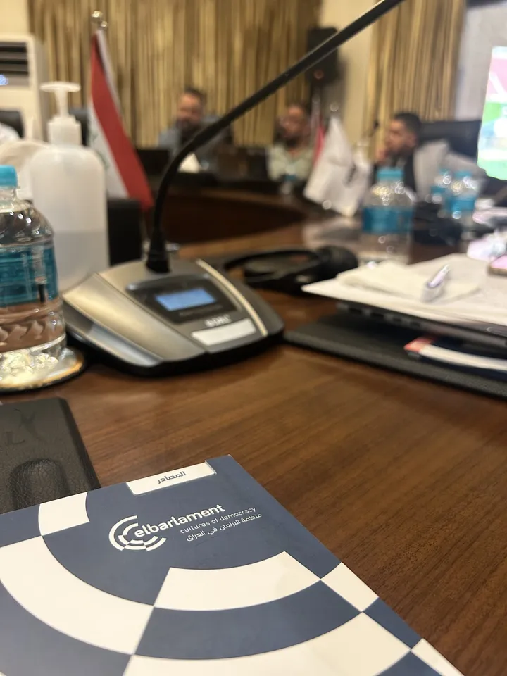 Close-up of a conference table with water bottles, a microphone and a folder reading "elbarlament", with people blurred in the background.