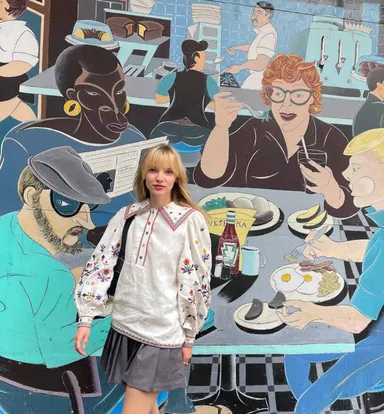 A woman stands in front of a colourful mural depicting people seated at a diner counter.