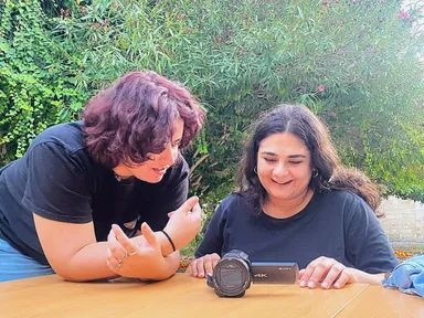 Two women lean over a table looking at a camcorder labeled "4K", with greenery in the background.
