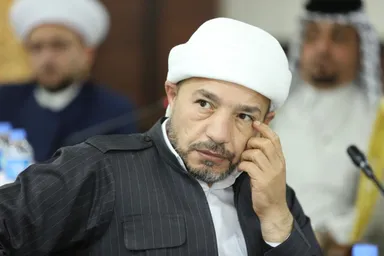 A man wearing a white turban touches his face while seated at a meeting, with two more men blurred in the background.