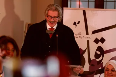 A man wearing glasses speaks at a podium with a microphone, in front of a banner reading "My Voice.".