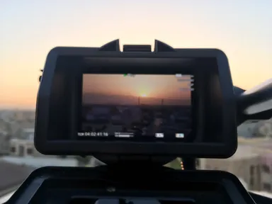 A camera's viewfinder screen shows a sunset over a city skyline with a timestamp overlay, mounted on a tripod against a hazy sky.