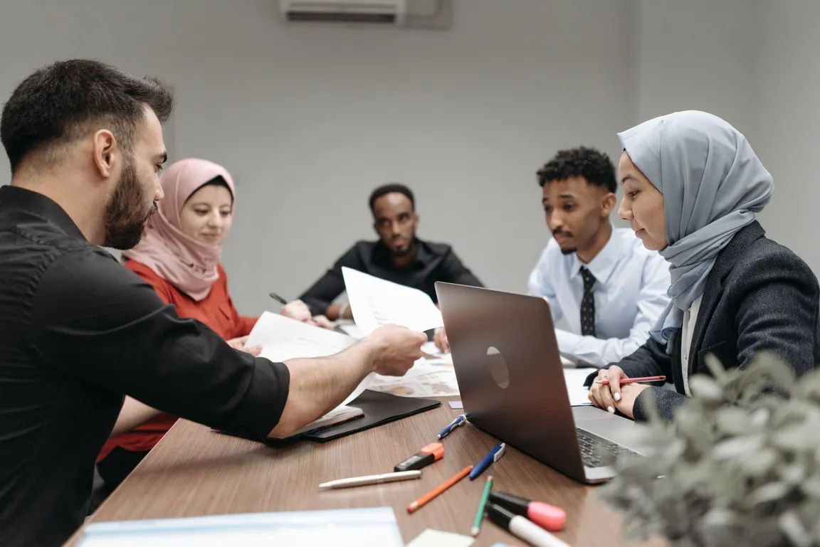 A group of people sit around a table with a laptop, papers and pens during a meeting, one person handing over a document.