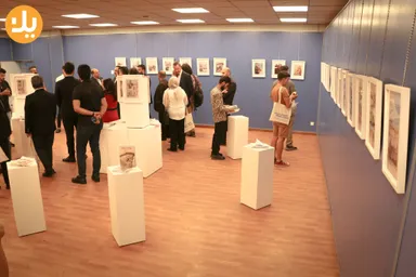 People browse framed pictures displayed on stands and a wall in an exhibition room, with a round orange logo in the corner and a tote bag printed "Mosul Tells" in the foreground.