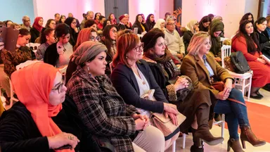 An audience of mostly women seated in rows in a room lit pink, listening attentively.