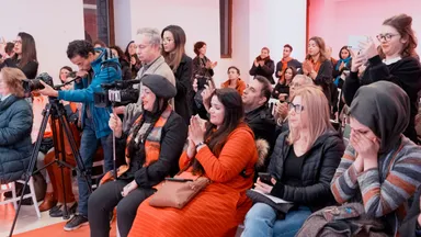 An audience applauds while a camera operator films the scene, several people wearing orange and one wiping her face.