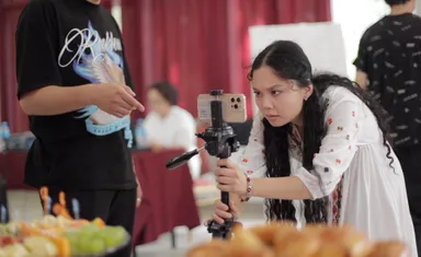 A woman adjusting a camera and phone mounted on a tripod near a table of food, with two other people standing nearby.
