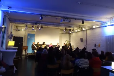 A wide view of a panel discussion on a stage under blue lighting, with a screen, a banner, a podium with a monitor, and a large seated audience in the foreground.