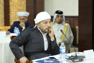 A man wearing a white turban rests his fingers near his chin at a conference table, with two more men blurred in the background.
