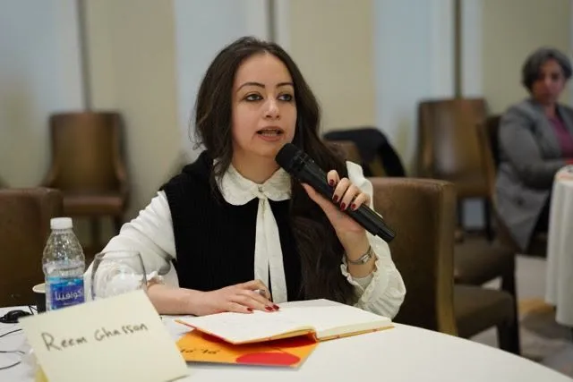 A woman speaks into a microphone while seated at a table with an open notebook, a water bottle, and a name card, another woman visible blurred in the background.