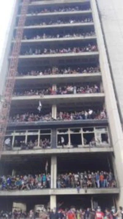 People fill every floor and balcony of a multi-story building overlooking a crowd gathered on the ground below.