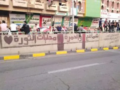 Arabic graffiti and a heart symbol cover a wall below a railing where people sit, with shop signs on the buildings behind them.
