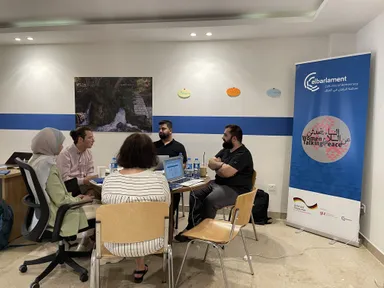 Five people sit in discussion with laptops beside a banner reading "elbarlament cultures of democracy" and "Women Talking Peace", a wall photo of a waterfall behind them.