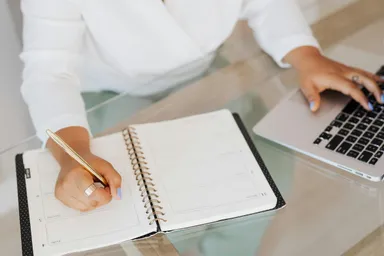 Close-up of a person's hands writing in an open notebook while typing on a laptop at a glass table.