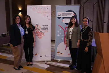 Four women stand together in front of two banners, one reading "WOMEN TAKING THE LEAD" with Arabic text and a hand-and-triangles logo, the other reading "Strengthening Participation, Peaceful Coexistence and Equality in Iraq" and "giz".