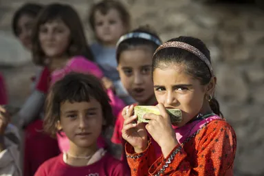 A girl eats a slice of fruit in front of several other children against a stone wall.