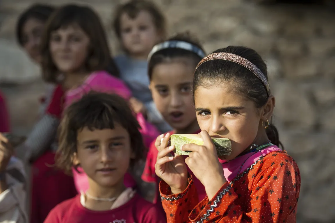 A girl eats a slice of fruit in front of several other children against a stone wall.