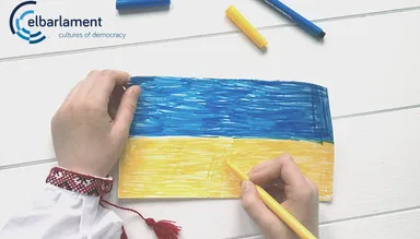 Two hands colour a paper Ukrainian flag in blue and yellow marker on a wooden table, with the elbarlament logo reading "elbarlament cultures of democracy" in the top left corner.