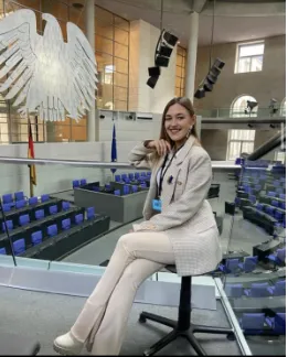 A woman sits smiling on a stool overlooking the empty plenary chamber of a parliament building, beneath a large stylised eagle emblem and blue seating rows.